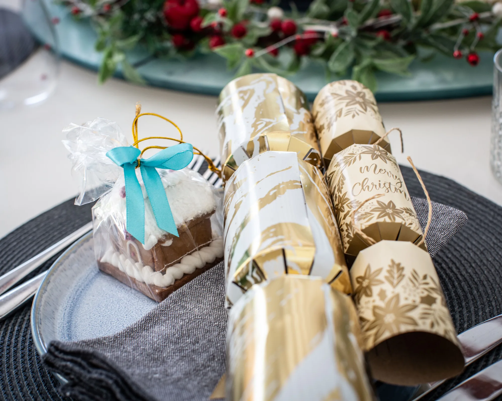 A Christmas table setting with gold and white crackers on a navy plate, a grey napkin and a small wrapped gift with a blue ribbon.