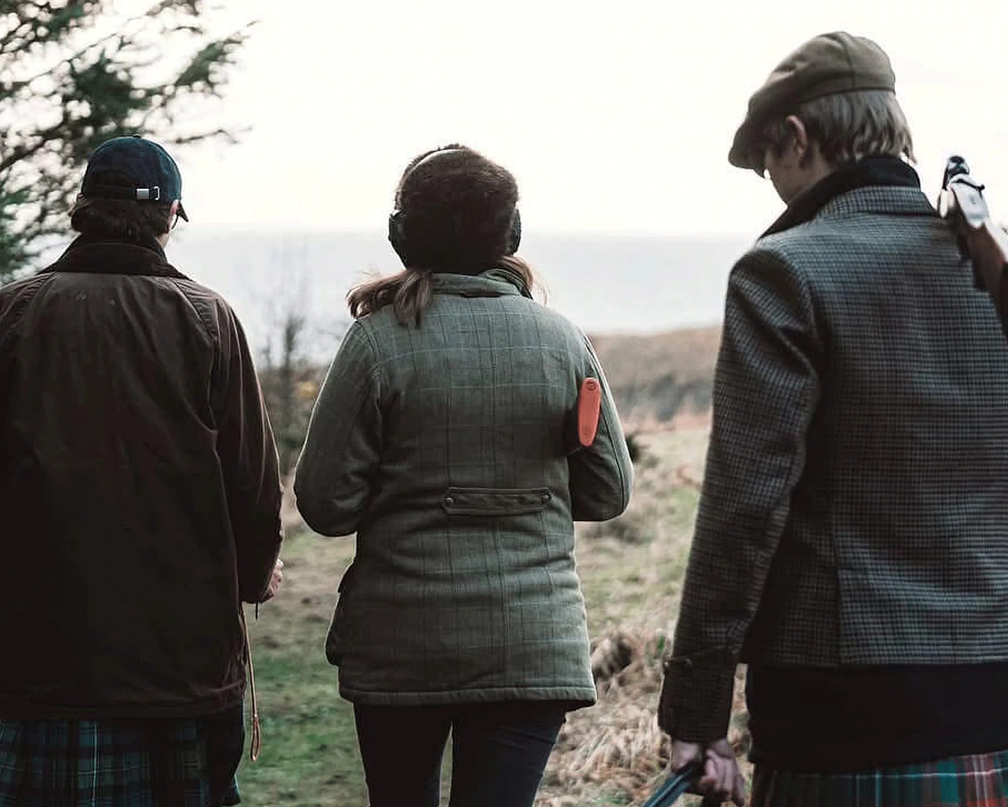 Three people in tweed and waxed jackets walking out towards the stands at Seal's Cove, one carrying a shotgun broken over his arm, the sea behind them.
