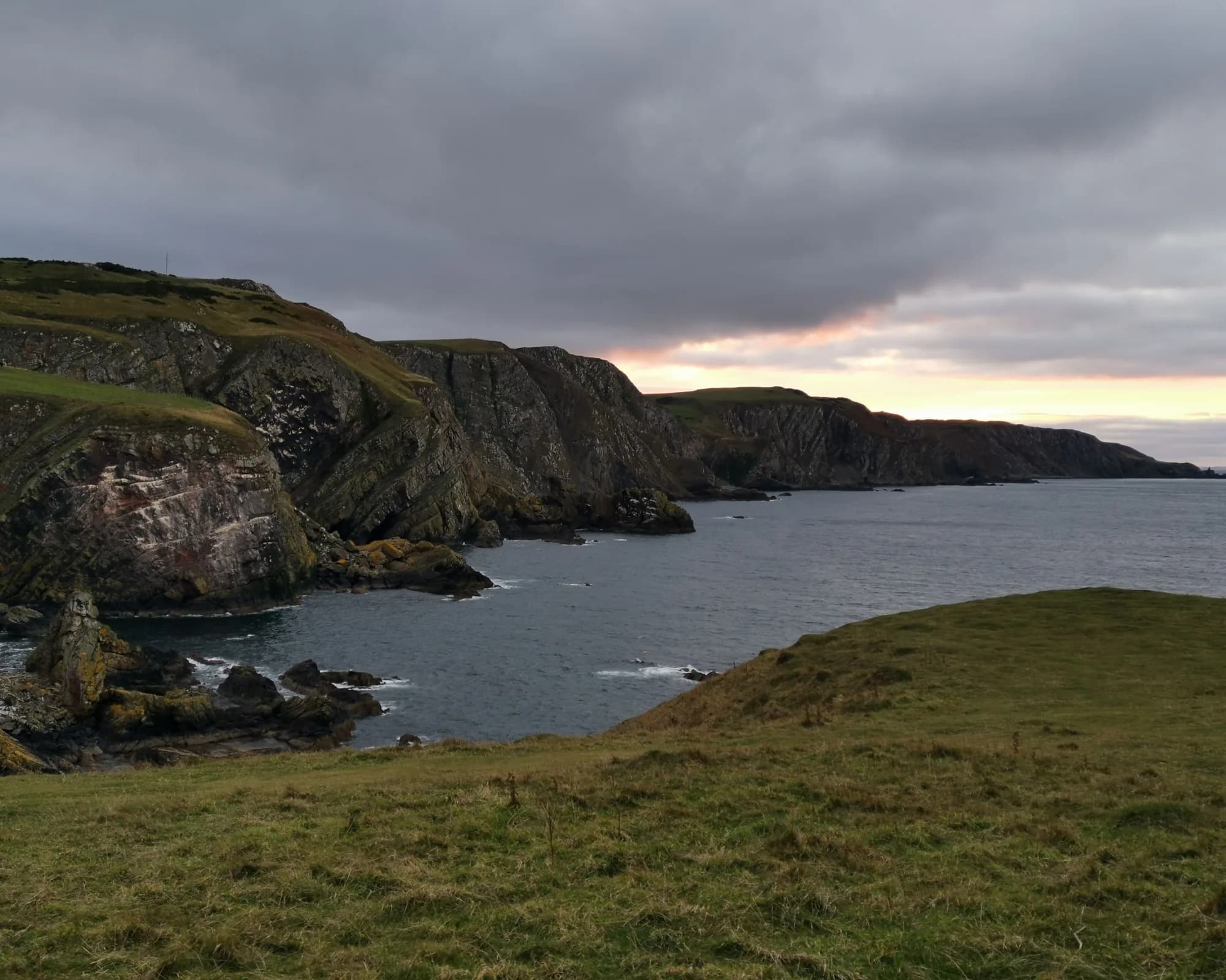 Eine grasbewachsene Klippe über einer felsigen Bucht an der schottischen Küste unter schwerem Abendhimmel. Ein Archivfoto der schottischen Küste, nicht von Cove Bay selbst.