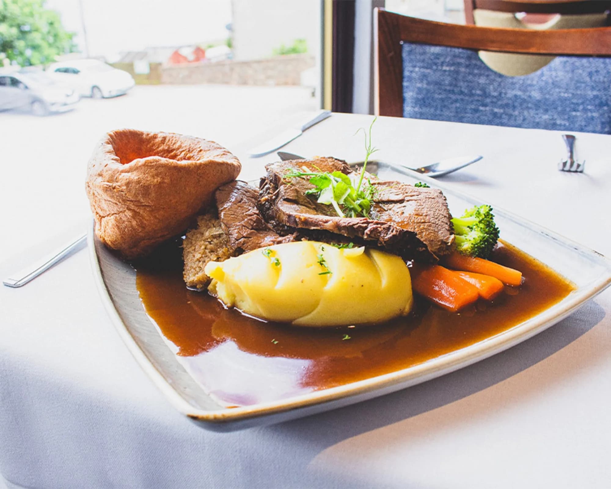 A plate of sliced roast beef with a Yorkshire pudding, mashed potato, carrots, broccoli and gravy, on a white tablecloth beside a restaurant window.